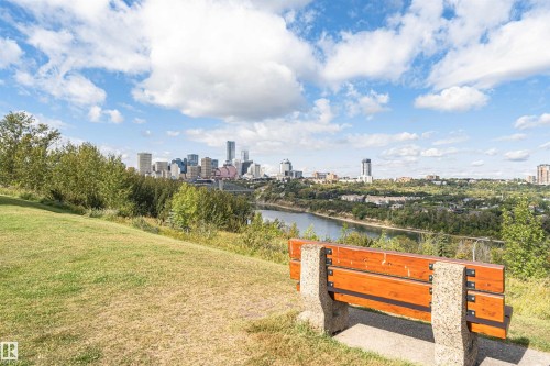 Expansive green space with a wood and stone park bench, offering a panoramic cityscape across the river - 8743 Strathearn Drive Nw, Edmonton, AB 