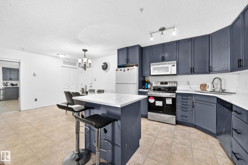 Modern kitchen featuring dark blue cabinetry, white countertops, stainless steel appliances, and a central island with seating - #133 16311 95 St Nw, Edmonton, AB 