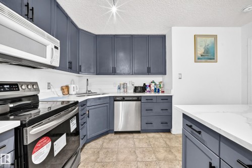 Blue cabinetry with matte black hardware, white countertops, built-in microwave, stainless steel dishwasher, and a stainless steel oven - #133 16311 95 St Nw, Edmonton, AB 