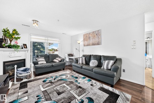 Spacious living area featuring wood-finish flooring, a white mantel fireplace, large windows, and recessed lighting - #133 16311 95 St Nw, Edmonton, AB 