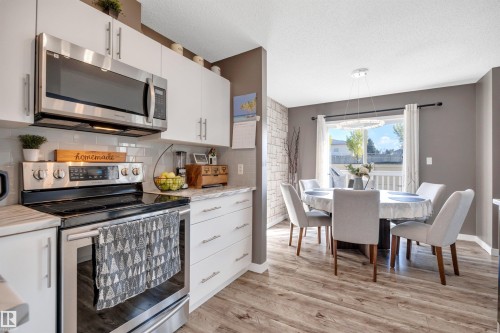Modern kitchen with stainless steel appliances, white cabinetry, a subway tile backsplash, and wood-finish flooring extending into the dining area with a contemporary light fixture - #13 6608 158 Av Nw, Edmonton, AB 
