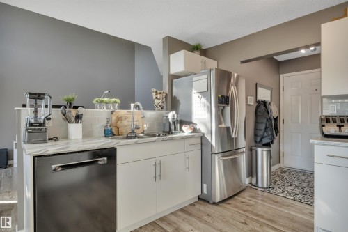 Kitchen with wood-finish flooring, white cabinetry, integrated stainless steel appliances, a tile backsplash, and a double basin sink - #13 6608 158 Av Nw, Edmonton, AB 