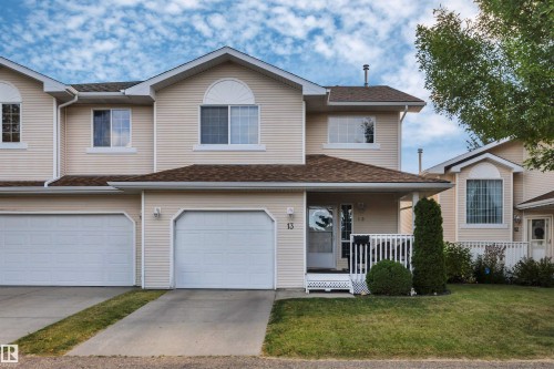 Cream-colored siding exterior, two attached single-car garages, white front porch railing, arched window accents, and a shingled roof - #13 6608 158 Av Nw, Edmonton, AB 