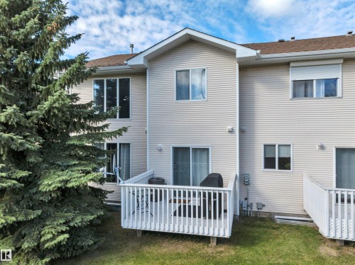 Rear exterior featuring light-colored siding, multiple windows, a private outdoor deck, and a sliding glass door - #13 6608 158 Av Nw, Edmonton, AB 