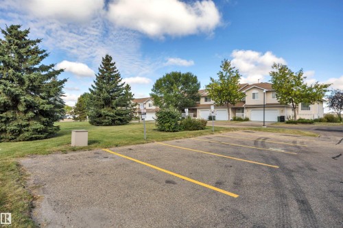Paved parking area with marked spaces, bordered by a landscaped green space with mature trees - #13 6608 158 Av Nw, Edmonton, AB 
