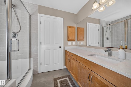 Built-in tub with glass enclosure, single vanity with light wood-finish cabinetry, integrated sink, and white countertop - 121 64 Street Sw, Edmonton, AB 