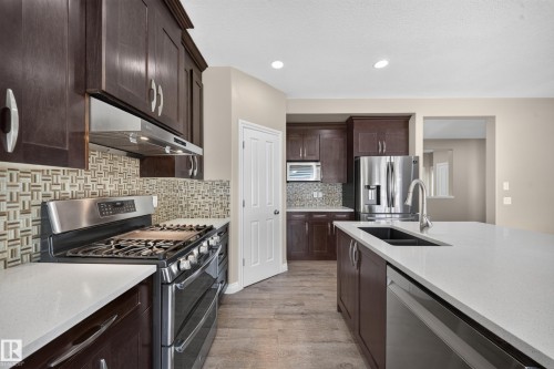 Kitchen featuring dark wood-finish cabinetry, stainless steel appliances, light-toned countertops, a mosaic tile backsplash, and wood-finish flooring - 121 64 Street Sw, Edmonton, AB 