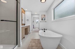 Contemporary bathroom featuring a freestanding soaking tub with matte black fixtures, a glass-enclosed shower, a wood-finish vanity with a white countertop, and light-colored floor tiles - 