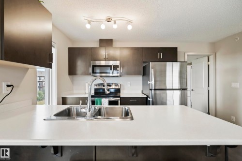 Kitchen featuring a large island with a stainless steel sink, dark wood-finish cabinetry, stainless steel appliances, and track lighting - #419 5515 7 Av Sw, Edmonton, AB 