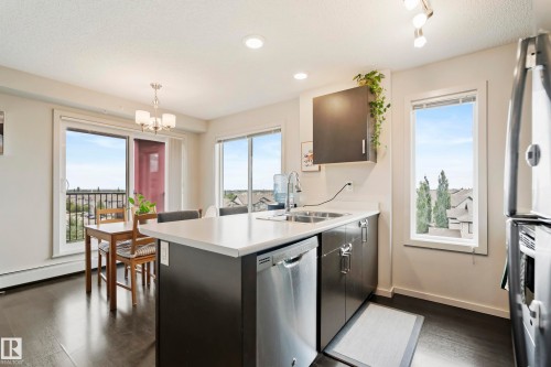 Kitchen featuring wood-finish flooring, stainless steel appliances, dark cabinetry, and an eat-in island with a white countertop - #419 5515 7 Av Sw, Edmonton, AB 