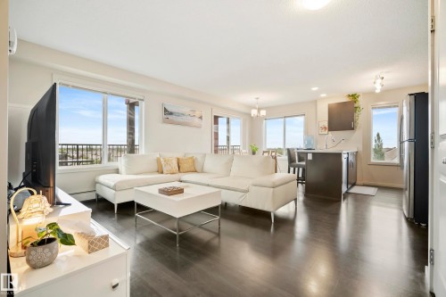 Open-concept living space featuring wood-finish flooring, large windows, a sliding glass door, kitchen island with seating, and stainless steel appliances - #419 5515 7 Av Sw, Edmonton, AB 