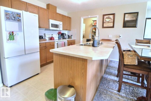 Kitchen featuring wood cabinetry, white appliances, light-toned countertops, and a kitchen island with an integrated sink - #421 592 Hooke Rd Nw, Edmonton, AB 