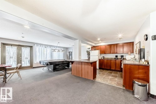 Spacious common room featuring carpet flooring, double glass panel doors, a three-light linear pendant, and a kitchen area with wood-finish cabinetry - #421 592 Hooke Rd Nw, Edmonton, AB 