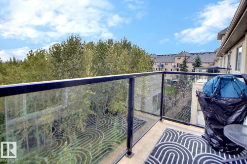Balcony with glass panel railing and metal frame, offering an elevated perspective of mature trees and surrounding residential structures - #421 592 Hooke Rd Nw, Edmonton, AB 
