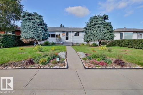 Front exterior with light blue siding, a central pathway, two mature evergreen trees, and landscaped garden beds - 13312 123A Street Nw, Edmonton, AB 