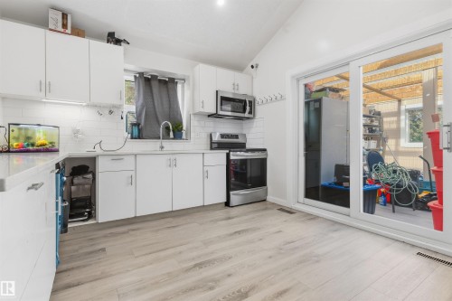 White cabinetry with silver hardware, light-toned wood-finish flooring, white subway tile backsplash, stainless steel appliances, and a sliding glass door - 4824 13 Avenue Nw, Edmonton, AB 