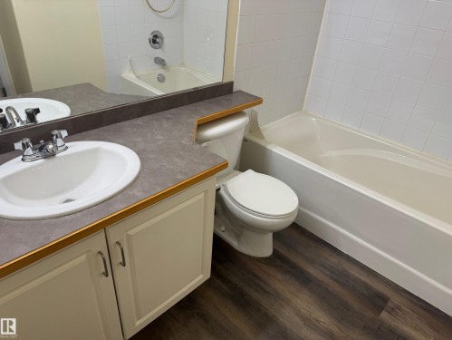 Bathroom featuring a built-in vanity with an integrated sink, a full-width mirror, a toilet, a bathtub with a tiled surround, and wood-finish flooring - #229 2305 35A Av Nw, Edmonton, AB 