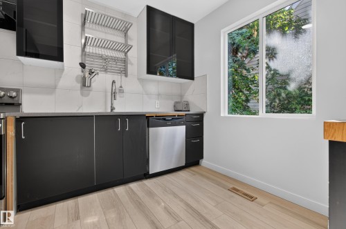Black cabinetry with integrated hardware, stainless steel dishwasher, light wood-finish flooring, and white window frame - 6807 112A Street Nw, Edmonton, AB 