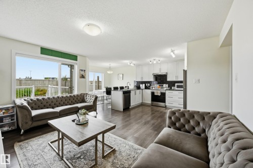Open-concept living area with dark wood-finish flooring, a contemporary kitchen featuring white cabinetry, dark backsplash, and stainless steel appliances, and a dining area with a modern chandelier - 941 Ebbers Crescent Nw, Edmonton, AB 