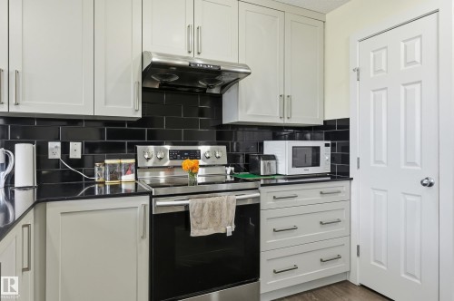 White shaker-style cabinetry with long pulls, black subway tile backsplash, stainless steel range with overhead hood, black countertops, and a white paneled door - 941 Ebbers Crescent Nw, Edmonton, AB 