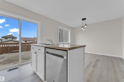 Kitchen island with integrated sink and stainless steel dishwasher, wood-finish flooring, sliding glass door to outdoor deck, pendant lighting, and a window with blinds - 
