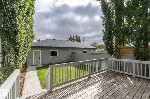 Expansive wood-finish deck with white railings, a concrete pathway, mature trees, and a gray siding structure featuring a dark shingle roof - 53/55 Michigan Street, Devon, AB 