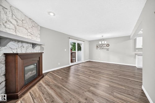 Wood-finish flooring, white stone-clad fireplace surround with wood mantel, built-in electric fireplace, sliding glass door, and a multi-light chandelier - 18146 81 Avenue Nw, Edmonton, AB 