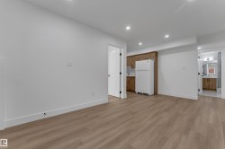 Recessed lighting illuminates wood-finish flooring, white walls, and a kitchen area featuring wood cabinetry, a white refrigerator, and a white tile backsplash - 