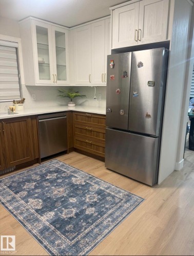 White upper cabinetry with glass-front accents, wood-finish lower cabinetry, light-toned wood-finish flooring, and a stainless steel refrigerator - 3830 22 Street Nw, Edmonton, AB 