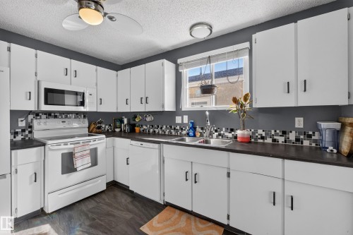 Kitchen featuring white cabinetry, dark countertops, a mosaic tile backsplash, stainless steel double basin sink, and wood-finish flooring - 333 Kirkpatrick Crescent Nw, Edmonton, AB 