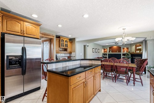 Open-concept kitchen and dining area featuring a kitchen island with black countertop, wood-finish cabinetry, stainless steel refrigerator, recessed lighting, and tiled flooring - 703 107A Street Sw, Edmonton, AB 