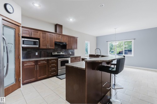 Open-concept kitchen featuring dark wood-finish cabinetry, stainless steel appliances, a stone-finish countertop island with seating, and tile flooring - 11608 96 Street Nw, Edmonton, AB 
