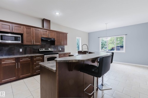 Kitchen featuring dark wood-finish cabinetry, stainless steel appliances, dark tile backsplash, and an island with an integrated sink - 11608 96 Street Nw, Edmonton, AB 