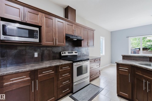 Kitchen featuring rich wood-finish cabinetry, stainless steel appliances, dark tile backsplash, and light tile flooring - 11608 96 Street Nw, Edmonton, AB 