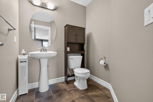 Pedestal sink with chrome fixtures, oval mirror, wall-mounted lighting, and brown floor tiling - 1850 Rutherford Road Sw, Edmonton, AB 