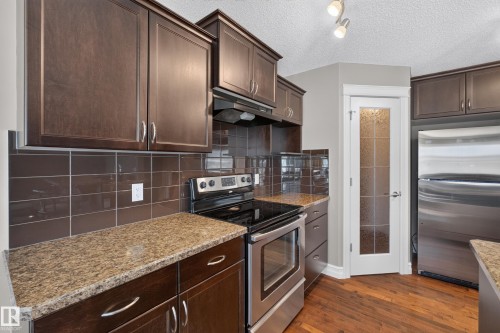 Kitchen featuring wood-finish cabinetry, speckled countertops, stainless steel appliances, a tile backsplash, and wood-finish flooring - 1850 Rutherford Road Sw, Edmonton, AB 