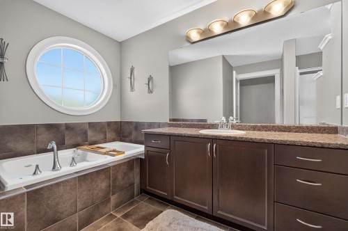 Built-in soaking tub with chrome fixtures, round window, tiled surround, and dual vanity with integrated sink and dark wood-finish cabinetry - 1850 Rutherford Road Sw, Edmonton, AB 