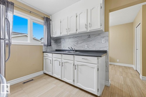 Full white cabinetry, dark countertop, marble-patterned backsplash, light-toned tiled flooring, and a large window - 11317 22 Avenue Nw, Edmonton, AB 