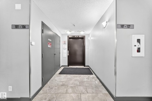 Corridor with large format tile flooring, wall-mounted light fixtures, and a dark-toned elevator door - #306 5370 Chappelle Rd Sw, Edmonton, AB 