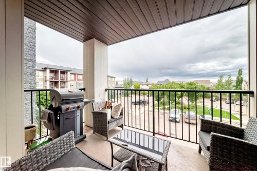 Covered balcony featuring a textured ceiling, a stone accent wall, and a black metal railing - #306 5370 Chappelle Rd Sw, Edmonton, AB 