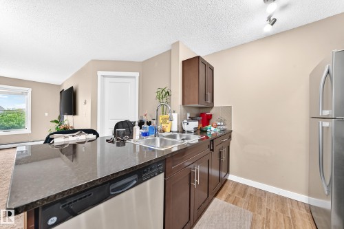 Kitchen featuring dark wood cabinetry, a double basin stainless steel sink, stainless steel dishwasher, and wood-finish flooring - #306 5370 Chappelle Rd Sw, Edmonton, AB 