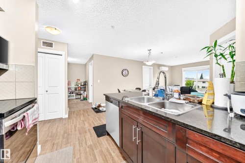 Kitchen featuring dark wood-finish cabinetry, a double basin stainless steel sink with a high-arc faucet, and dark countertops - #306 5370 Chappelle Rd Sw, Edmonton, AB 