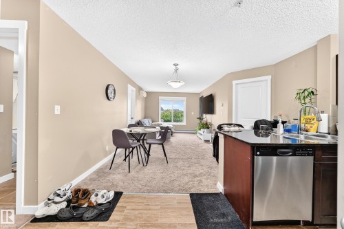 Open concept living area with carpeted flooring, kitchen island with dark countertop, stainless steel dishwasher, and a window with exterior scenery - #306 5370 Chappelle Rd Sw, Edmonton, AB 