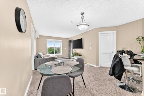 Open-concept living area with carpet flooring, a large window, a ceiling-mounted light fixture, and a white paneled door - #306 5370 Chappelle Rd Sw, Edmonton, AB 