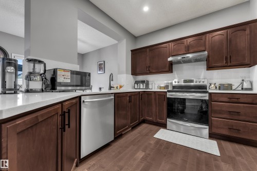 Hardwood-finish flooring, L-shaped white countertop, stainless steel appliances, dark wood cabinetry, and a white tile backsplash - 3230 21A Avenue Nw, Edmonton, AB 