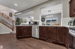 Rich wood-finish flooring, dark wood cabinetry, light countertops, stainless steel dishwasher, and a staircase with wood newel posts and black metal spindles - 