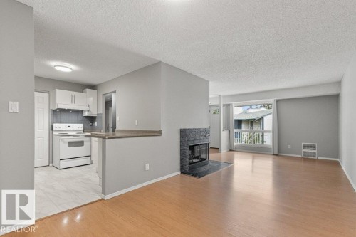 Wood-finish flooring, a contemporary fireplace with a dark stone surround, white kitchen cabinetry, a tile kitchen backsplash, and a breakfast bar - #4 14620 26 St Nw, Edmonton, AB 