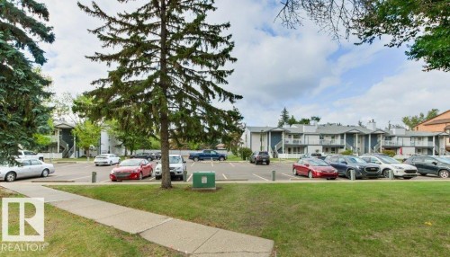 Paved parking area surrounded by mature trees, featuring multiple residential buildings with balconies and exterior staircases - #4 14620 26 St Nw, Edmonton, AB 