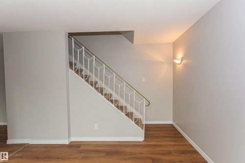 Staircase featuring a white wrought iron railing with a brass-finish handrail, wood-finish flooring, light grey wall paint, white baseboards, and a wall-mounted sconce - Edmonton, AB - Indoor Photo Showing Other Room