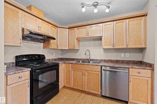 Kitchen featuring wood-finish flooring, light wood cabinetry, a double basin stainless steel sink, a stainless steel dishwasher, and a black electric range with an overhead vent - 15504 102 Avenue Nw, Edmonton, AB 